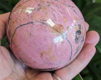May include: A pink and gold sphere of polished rhodochrosite, a type of gemstone, with black veins running through it.