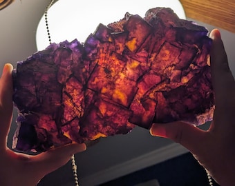 Puede incluir: Un gran cristal de fluorita púrpura y naranja con una textura rugosa y natural. El cristal es translúcido y tiene una variedad de colores y patrones.