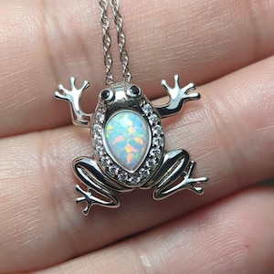 May include: Silver-toned frog pendant necklace. The frog pendant features an oval, iridescent opal-like stone, surrounded by small clear stones. The frog's body and legs are detailed with a textured finish, and the necklace chain is delicate.