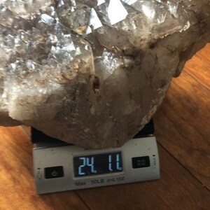 May include: A large, clear quartz crystal cluster weighing 24.11 ounces. The cluster is on a digital scale with a white background.