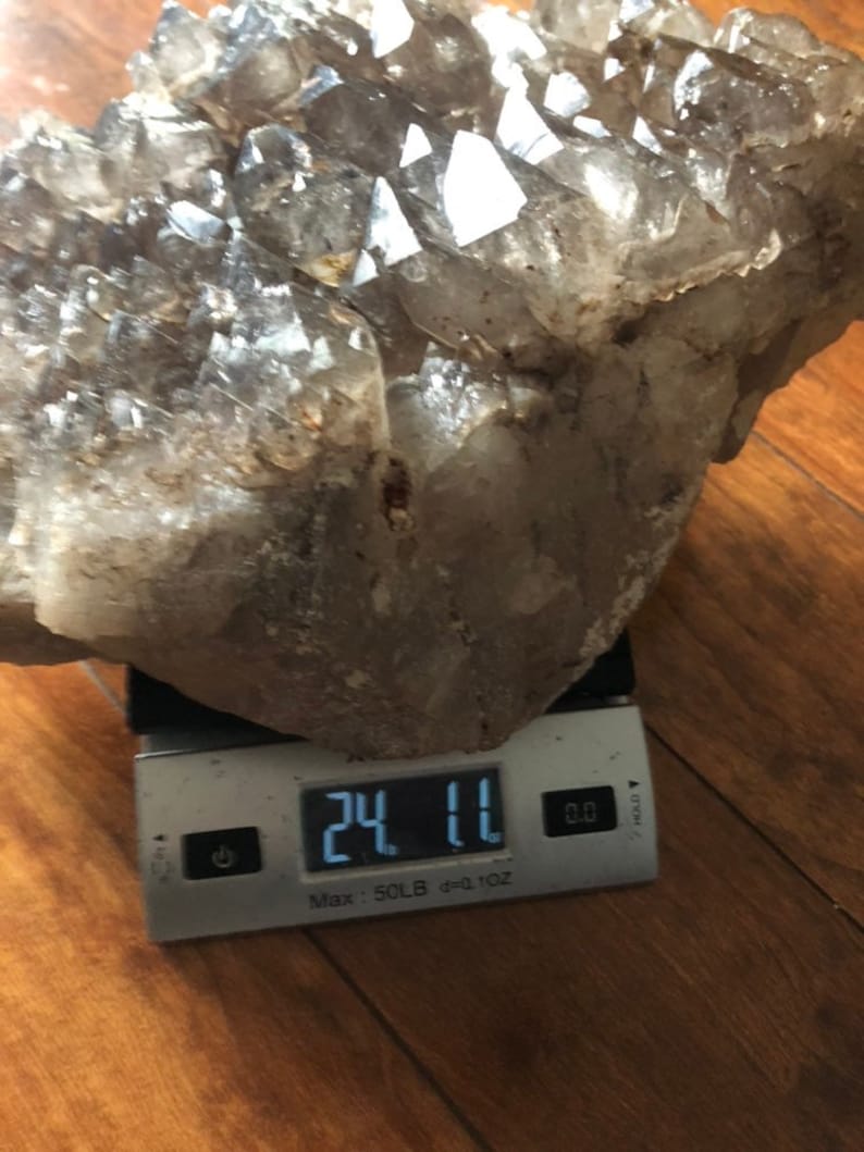 May include: A large, clear quartz crystal cluster weighing 24.11 ounces. The cluster is on a digital scale with a white background.