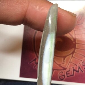 May include: A translucent white oval-shaped cabochon with a green and white inclusion. The cabochon is held by a finger.