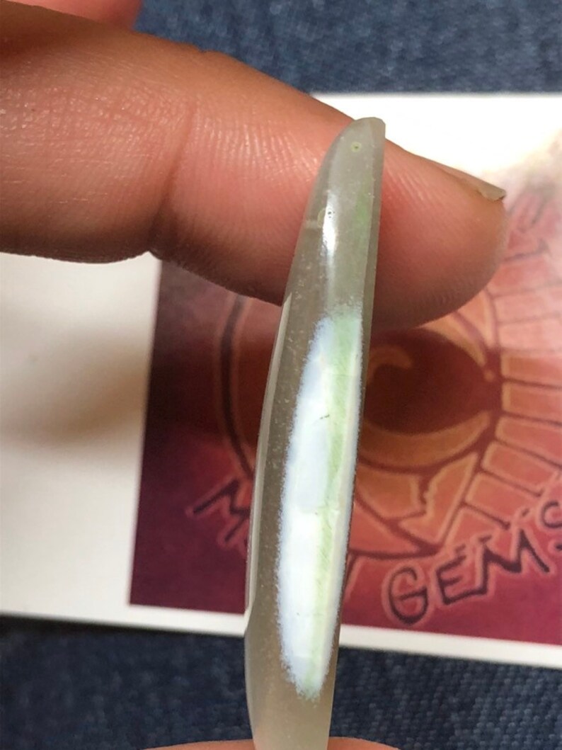 May include: A translucent white oval-shaped cabochon with a green and white inclusion. The cabochon is held by a finger.
