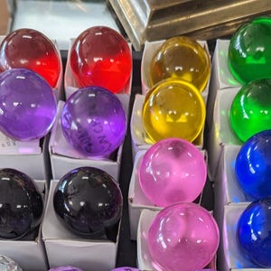 May include: A collection of 12 glass spheres in various colors, including red, purple, yellow, green, pink, and blue. The spheres are displayed in a row on a white surface.