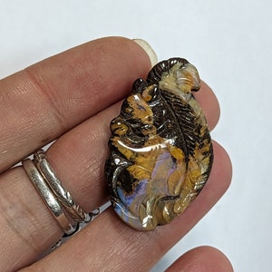 Puede incluir: Un colgante de ópalo tallado en forma de hoja con colores iridiscentes marrones, amarillos y azules.