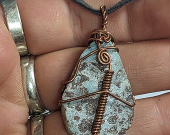 Pode incluir: Um pingente em forma de lágrima com uma pedra azul e marrom envolvida em fio de cobre. A pedra tem um padrão salpicado.