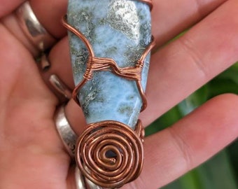 Pode incluir: Um pingente de pedra preciosa azul claro e verde envolto em fio de cobre. O pingente tem a forma de uma lágrima e tem um design em espiral na parte inferior.