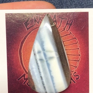May include: A teardrop-shaped cabochon of white, gray, and brown opal. The opal is set on a card with the text "EMPATH" and "MOONS" visible.