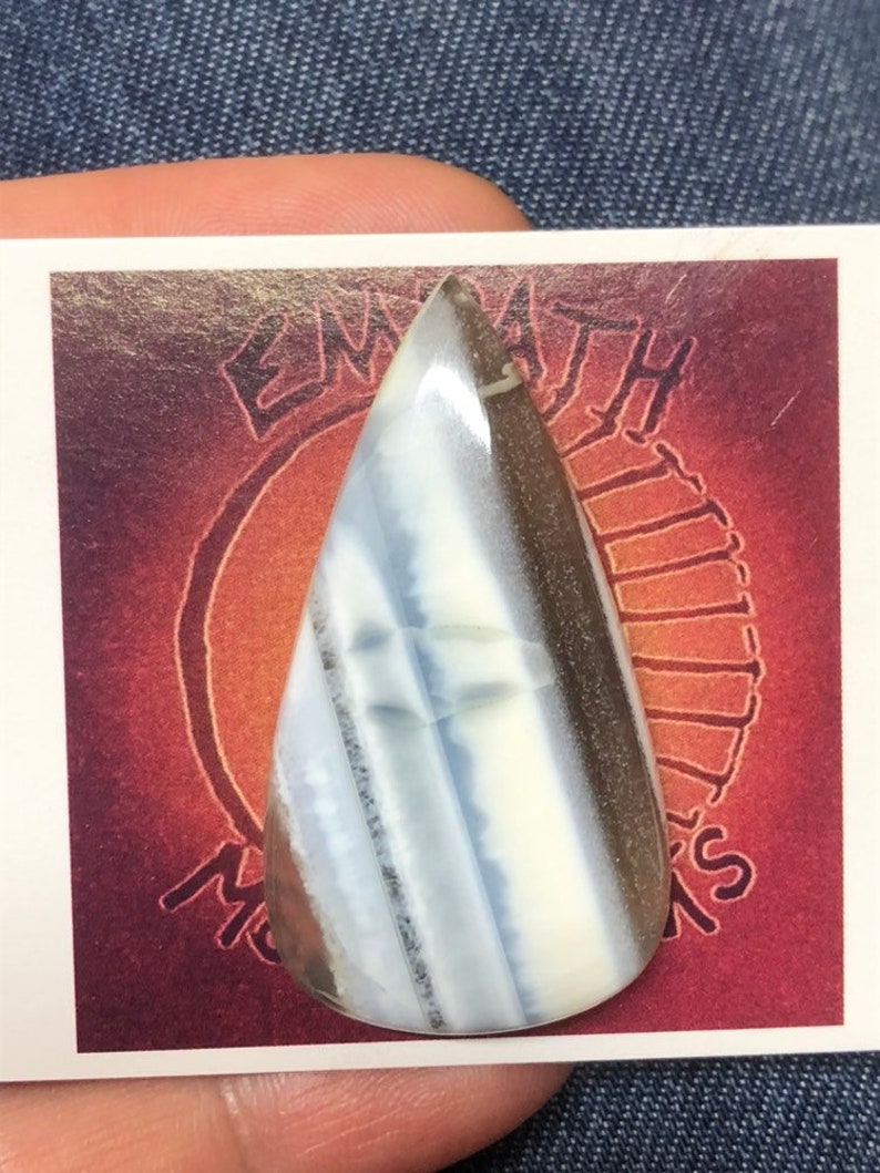May include: A teardrop-shaped cabochon of white, gray, and brown opal. The opal is set on a card with the text "EMPATH" and "MOONS" visible.
