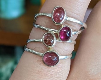 May include: Four silver rings with pink and red oval gemstones. The rings are stacked on a finger.