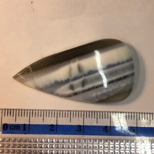 May include: A white, gray, and black banded agate cabochon with a teardrop shape. The cabochon is approximately 4 cm long.