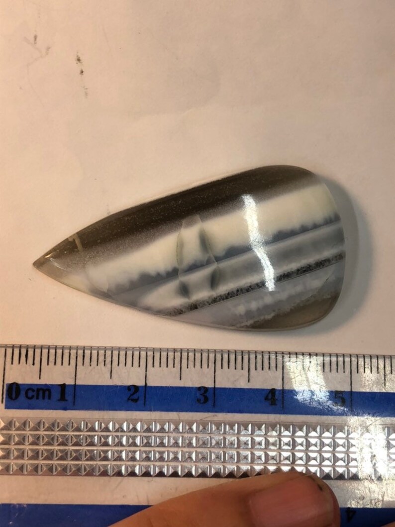May include: A white, gray, and black banded agate cabochon with a teardrop shape. The cabochon is approximately 4 cm long.