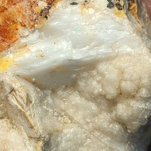 May include: A close-up of a white opal with a rough, natural surface. The opal has a milky white color with a hint of iridescent blue. The opal is surrounded by a matrix of white and brown rock.