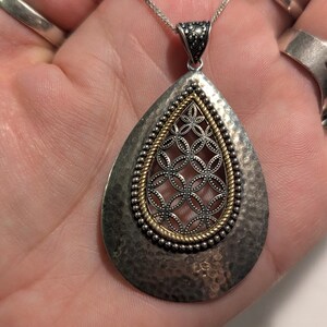 May include: A silver and gold teardrop-shaped pendant necklace with a delicate filigree design. The pendant has a hammered texture and is accented with small beads.
