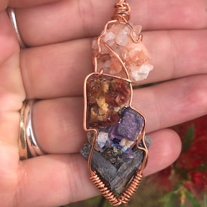 May include: A copper wire wrapped pendant featuring a variety of crystals, including a cluster of pink selenite, a piece of red carnelian, a piece of amethyst, and a piece of black kyanite.