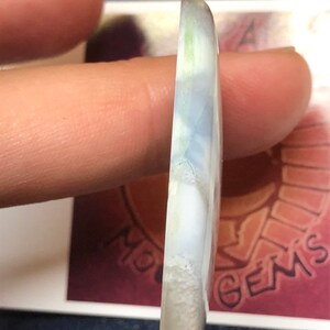 May include: A white cabochon gemstone with blue and green inclusions. The gemstone is smooth and polished, with a rounded shape.