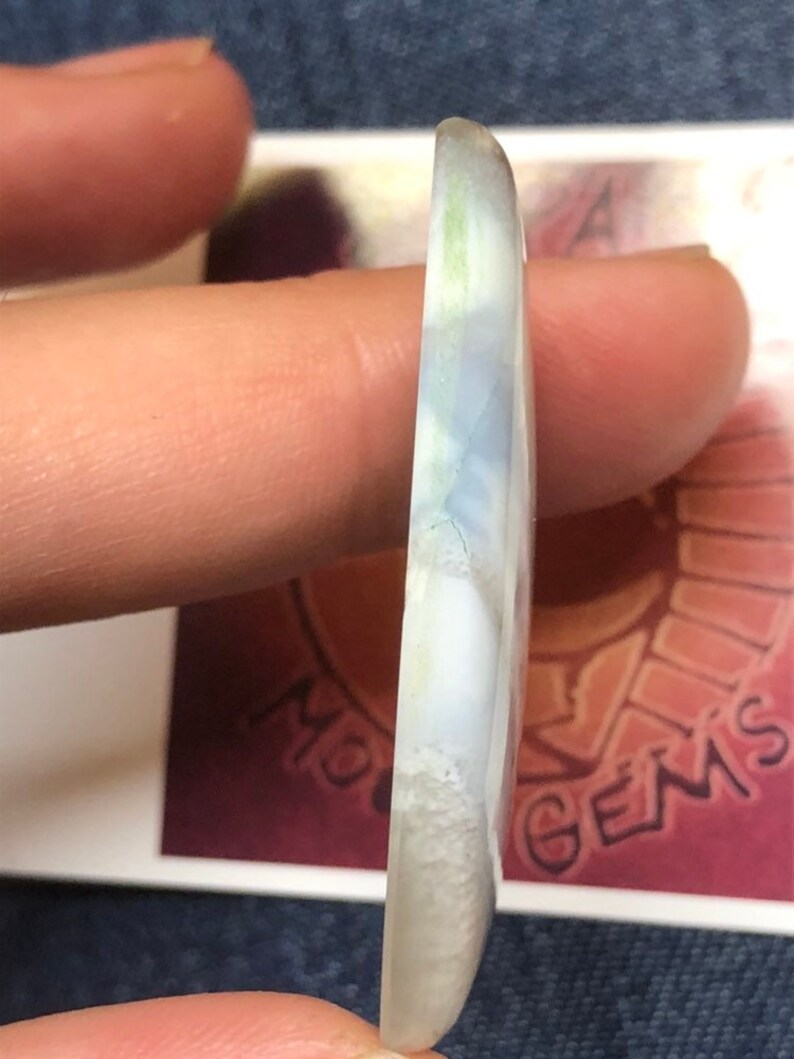 May include: A white cabochon gemstone with blue and green inclusions. The gemstone is smooth and polished, with a rounded shape.