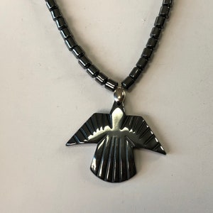 May include: A black hematite necklace with a pendant shaped like a stylized bird. The pendant has a shiny, polished finish.
