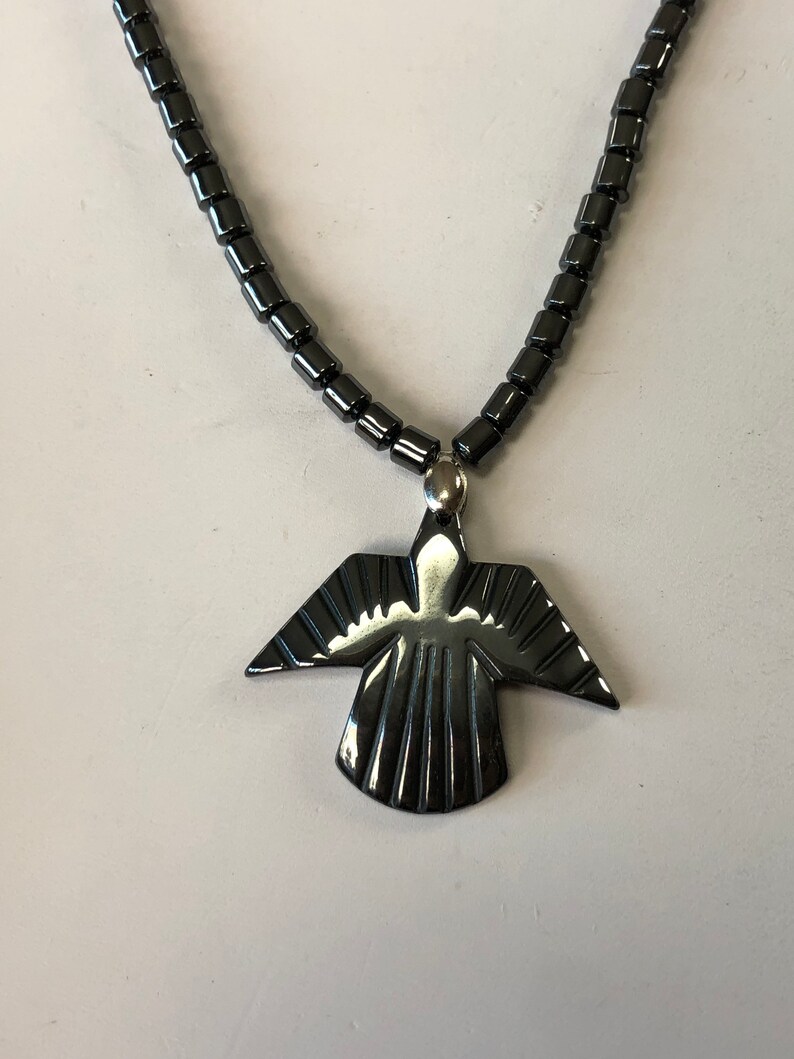 May include: A black hematite necklace with a pendant shaped like a stylized bird. The pendant has a shiny, polished finish.