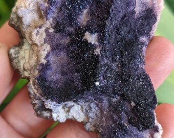 May include: A close-up of a purple and grey fluorite crystal with black inclusions. The crystal is rough and has a natural, unpolished look.