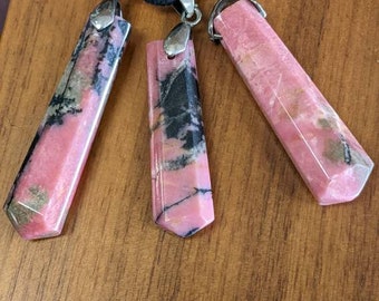 May include: Three pink and black gemstone pendants on a black cord necklace. The pendants are faceted and have a silver tone bail.