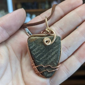 Puede incluir: Un collar de cordón de cuero marrón con un colgante envuelto en alambre de cobre que presenta una piedra gris y marrón.