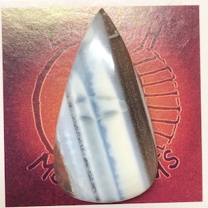 May include: A teardrop-shaped cabochon of white, blue, and brown banded opal. The opal is polished and has a smooth, glossy finish.