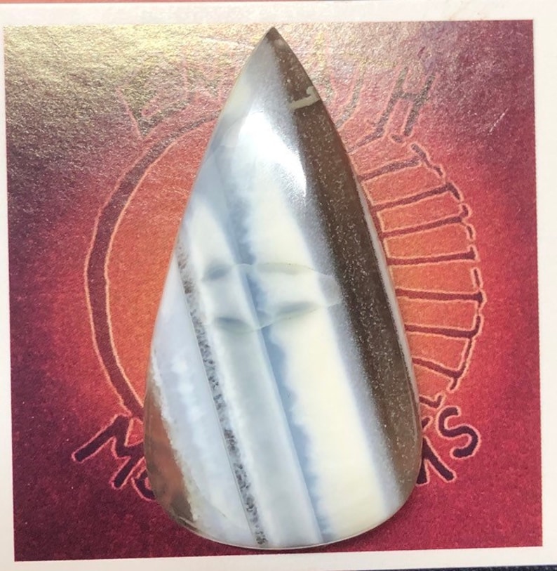 May include: A teardrop-shaped cabochon of white, blue, and brown banded opal. The opal is polished and has a smooth, glossy finish.