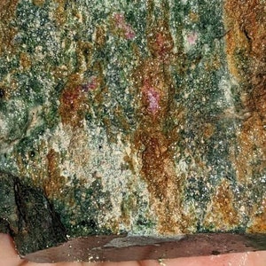 May include: A close-up of a rough, green, brown, and white rock with small, pink crystals embedded in it.