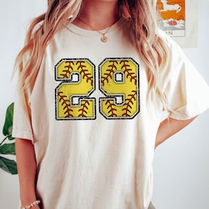 May include: A white t-shirt with a distressed, black outlined number 29. The numbers are filled with a yellow softball design with red stitching.