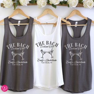 May include: Three gray tank tops with white text that reads "THE BACH Exclusive Club" and a graphic of two martini glasses. The text below the graphic reads "Emily's Bachelorette LAS VEGAS, NV". The middle tank top is white.