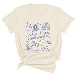 K&ouml;nnte beinhalten: Cremefarbenes T-Shirt mit blauem Grafikdesign. Das Design umfasst B&auml;ume, ein Lagerfeuer, eine H&uuml;tte und die Worte "Cabin Crew HAYWARD, WI 2025". Zus&auml;tzliche Illustrationen sind ein Waschb&auml;r, eine Angelrute, Fische, ein Stuhl und ein Kanu.