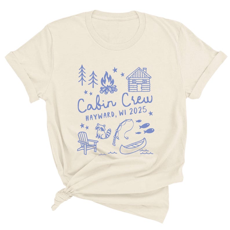 K&ouml;nnte beinhalten: Cremefarbenes T-Shirt mit blauem Grafikdesign. Das Design umfasst B&auml;ume, ein Lagerfeuer, eine H&uuml;tte und die Worte "Cabin Crew HAYWARD, WI 2025". Zus&auml;tzliche Illustrationen sind ein Waschb&auml;r, eine Angelrute, Fische, ein Stuhl und ein Kanu.