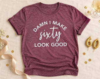 May include: A maroon short-sleeved t-shirt with white text that reads DAMN I MAKE sixty LOOK GOOD. The shirt features a crew neckline and a heathered fabric texture, presented as a flat lay garment.