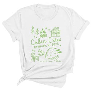 K&ouml;nnte beinhalten: Wei&szlig;es T-Shirt mit einem gr&uuml;nen Grafikdesign. Das Design enth&auml;lt den Text "Cabin Crew HAYWARD, WI 2025" sowie Illustrationen einer H&uuml;tte, B&auml;ume, ein Lagerfeuer, ein Waschb&auml;r, eine Angelrute, Fische, ein Kanu und einen Stuhl.