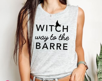 May include: A light gray sleeveless muscle tank top featuring the black text "WITCH way to the BARRE" with a small black witch hat icon above the word "WITCH". The shirt has a crew neckline and is worn tucked into blue denim jeans.