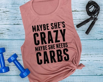 May include: A dusty rose muscle tank top with the text "MAYBE SHE'S CRAZY MAYBE SHE NEEDS CARBS" in black. Two blue dumbbells and a black jump rope are also in the image. The tank top is knotted at the bottom.