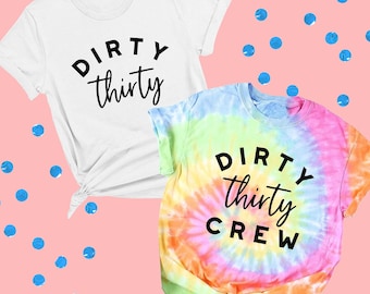 Puede incluir: Una camiseta blanca con el texto negro DIRTY thirty y una camiseta tie-dye multicolor con el texto negro DIRTY thirty CREW. Ambas camisetas tienen mangas cortas, cuello redondo y un detalle de dobladillo anudado en el lateral.