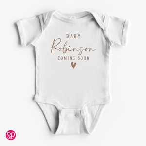 May include: White baby onesie with the text "BABY Robinson COMING SOON" in brown script and a brown heart graphic. The onesie has short sleeves and a snap closure.