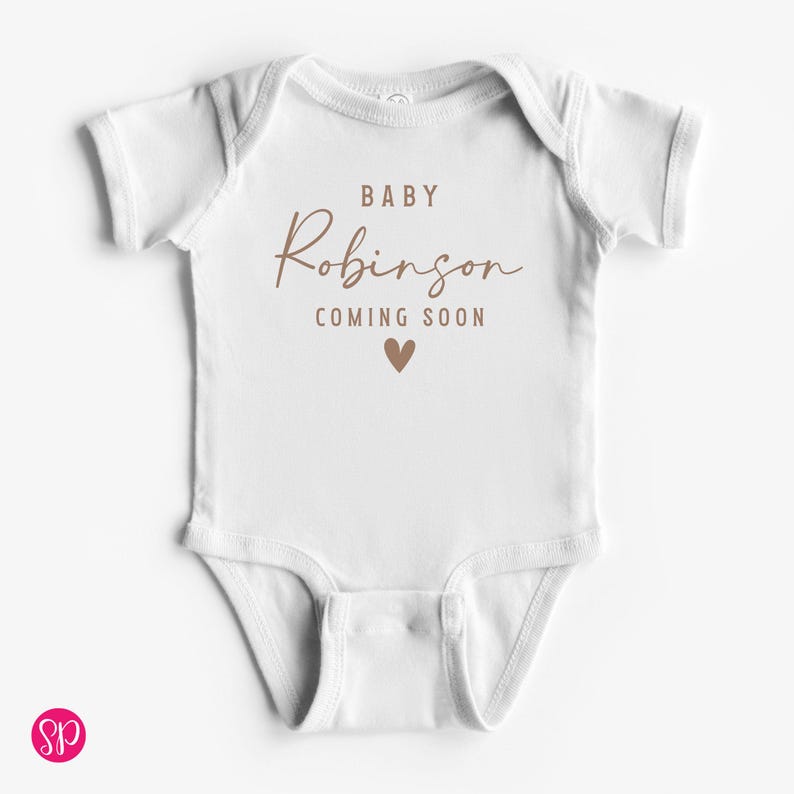 May include: White baby onesie with the text "BABY Robinson COMING SOON" in brown script and a brown heart graphic. The onesie has short sleeves and a snap closure.