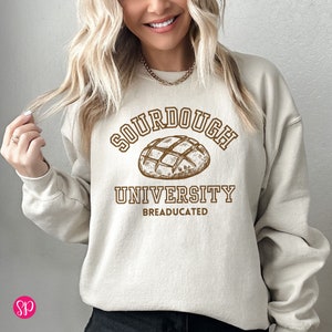 May include: A beige sweatshirt with a brown graphic of a sourdough loaf of bread. The graphic says "Sourdough University" and "BREADUCATED" underneath.
