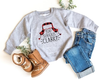 May include: A light gray long-sleeve crewneck sweatshirt featuring a red and black buffalo plaid trapper hat graphic above the black text "YOU SERIOUS CLARK?", paired with blue denim jeans and tan suede boots with fringe and faux fur lining.