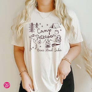 May include: Cream-colored t-shirt with a graphic design featuring camping-themed illustrations and text. The design includes trees, a tent, a campfire, and the words "Camp Peterson Bear Head Lake 2025". The shirt has short sleeves.