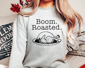 Puede incluir: Una sudadera gris claro de cuello redondo con el texto negro Boom. Roasted. sobre un dibujo lineal negro de un pavo asado en una bandeja. La prenda tiene mangas largas y un cuello acanalado, ofreciendo un aspecto sencillo y casual.
