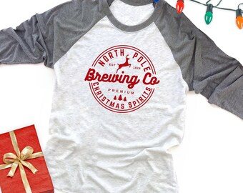 May include: A heather gray and white raglan baseball shirt featuring a red circular graphic on the chest. The text reads North Pole Brewing Co. Est 1824 Premium Christmas Spirits, accompanied by a reindeer and small pine tree icons.