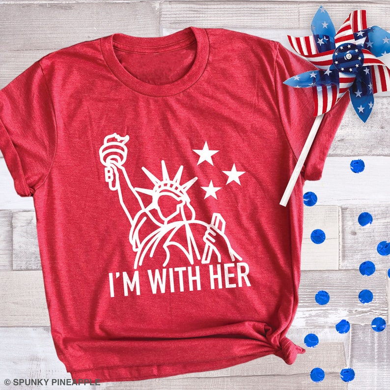 Puede incluir: Camiseta roja con un contorno blanco de la Estatua de la Libertad y el texto "I'm With Her".