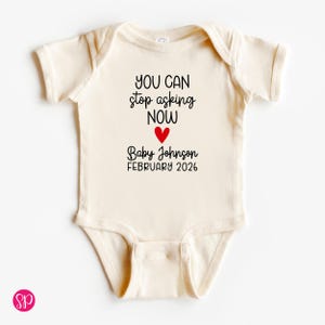 May include: Cream-colored baby onesie with the text "YOU CAN stop asking NOW" in black, a red heart, and "Baby Johnson FEBRUARY 2026". The onesie has short sleeves and a snap closure.
