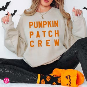 May include: A beige crewneck sweatshirt featuring the orange text PUMPKIN PATCH CREW printed on the front. The garment is paired with black leggings and orange fuzzy slippers with a jack-o'-lantern face design on the front.
