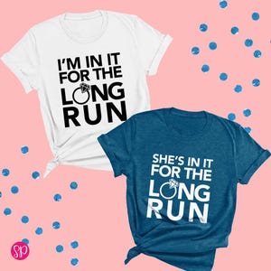 May include: Two t-shirts with a white background and a blue background. The white t-shirt has black text that says "I'm in it for the long run" with a diamond ring graphic. The blue t-shirt has white text that says "She's in it for the long run" with a diamond ring graphic.