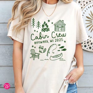 K&ouml;nnte beinhalten: Beiges T-Shirt mit gr&uuml;nem Aufdruck "Cabin Crew Hayward, WI 2025". Das Design zeigt B&auml;ume, ein Lagerfeuer, eine H&uuml;tte, einen Waschb&auml;ren, Angelausr&uuml;stung, ein Kanu und Fische.